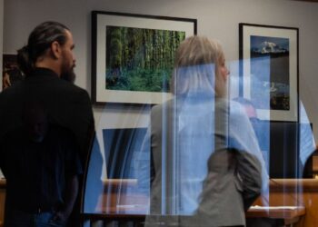 King County Sheriff’s Deputy Ricardo Arturo Cueva, a man with light-toned skin, a beard and hair in a ponytail, wears a suit, and stands facing away from the camera. To his right stands a woman with light-toned skin and gray, medium-length hair, wearing a gray suit jacket.