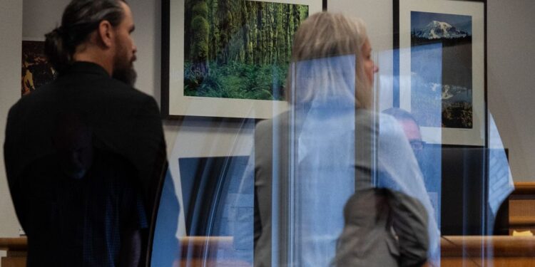 King County Sheriff’s Deputy Ricardo Arturo Cueva, a man with light-toned skin, a beard and hair in a ponytail, wears a suit, and stands facing away from the camera. To his right stands a woman with light-toned skin and gray, medium-length hair, wearing a gray suit jacket.