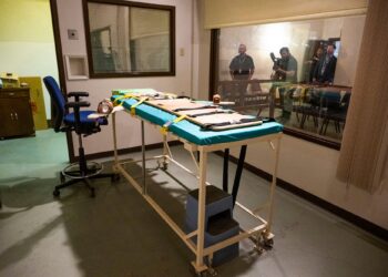 A photo shows a gurney and a blue chair inside a death chamber. An adjacent room is shown through a window, with three people standing and one person holding a camera inside.
