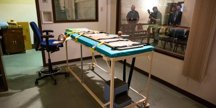 A photo shows a gurney and a blue chair inside a death chamber. An adjacent room is shown through a window, with three people standing and one person holding a camera inside.