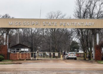 A photo shows a silver car going through a prison checkpoint entry with a large sign that reads “Mississippi State Penitentiary.” Bare trees are visible in the background beyond the entrance.