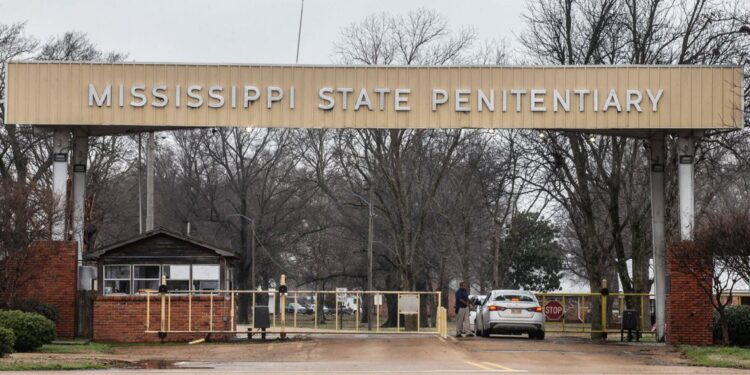 A photo shows a silver car going through a prison checkpoint entry with a large sign that reads “Mississippi State Penitentiary.” Bare trees are visible in the background beyond the entrance.