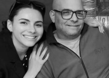 A black-and-white photo of a White woman wearing sunglasses on her head with her arm around her father, a bald White man who is wearing glasses.