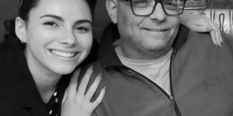 A black-and-white photo of a White woman wearing sunglasses on her head with her arm around her father, a bald White man who is wearing glasses.