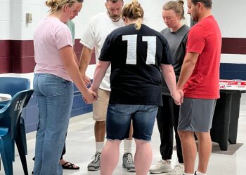 A group of six people hold hands with their heads facing downward. They are standing in a circle, inside a prison visitation room.