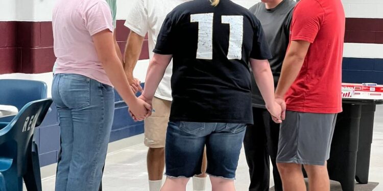 A group of six people hold hands with their heads facing downward. They are standing in a circle, inside a prison visitation room.