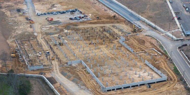 An aerial photo shows a site in the early stages of construction. Cars are parked in a lot near the site.