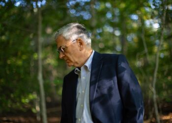 A photo shows an older White man with gray hair and glasses looking down while standing in a forest. The man is wearing a navy blue blazer and a blue button-down shirt.