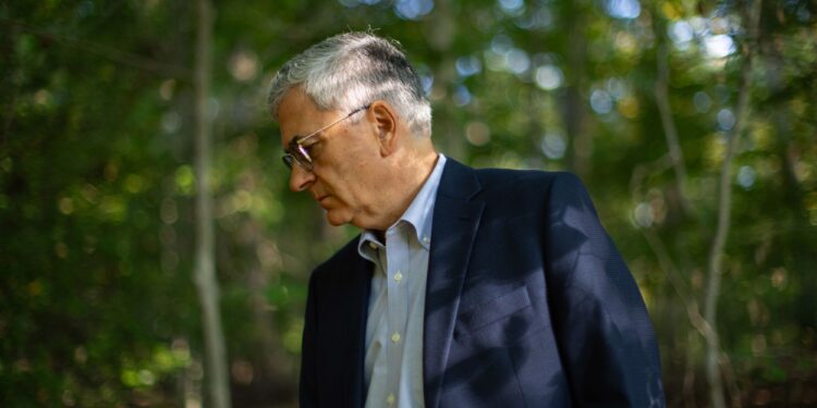 A photo shows an older White man with gray hair and glasses looking down while standing in a forest. The man is wearing a navy blue blazer and a blue button-down shirt.
