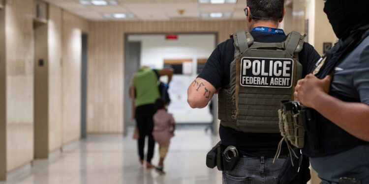 A photo shows the back of a light-skinned officer wearing glasses, a black T-shirt, gray jeans and a vest with the words "police federal agent." One police officer stands next to them. A person and a child walk down a hallway standing in the background.