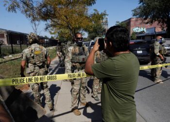 A photo shows a man with a medium skin tone and a olive green t-shirt holding his phone up toward a group law enforcement agents, who are standing in uniform on the other side of yellow police tape.