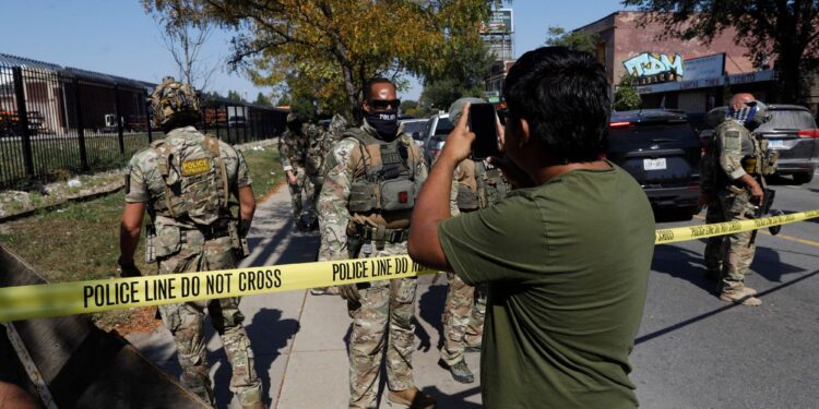 A photo shows a man with a medium skin tone and a olive green t-shirt holding his phone up toward a group law enforcement agents, who are standing in uniform on the other side of yellow police tape.