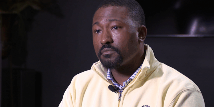 A Black man with a goatee wears a white collared shirt with blue stripes and a yellow jacket with a green logo.
