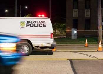 A photo shows a Cleveland Police van on a street at night parked by traffic cones, while a blue car starts to drive by from the left side of the photo.