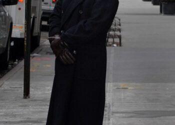 Jeffrey Epstein stands on a sidewalk, wearing a long dark coat, jeans, and brown gloves.