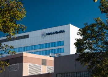 A photo of the MetroHealth Medical Center. The building is white and rectangular, has two lines of visible windows, and the word “MetroHealth” is on the top right of the building. In front of that building is a lower beige/orange-toned building.