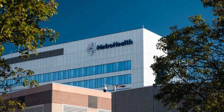 A photo of the MetroHealth Medical Center. The building is white and rectangular, has two lines of visible windows, and the word “MetroHealth” is on the top right of the building. In front of that building is a lower beige/orange-toned building.