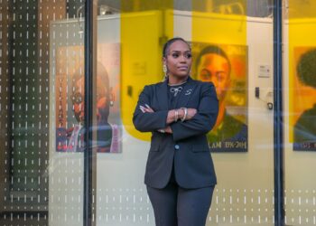 A photo of Shamari Jackson, a Black woman wearing a suit, standing outside of a building with glass windows. In the building space are paintings of homicide victims.