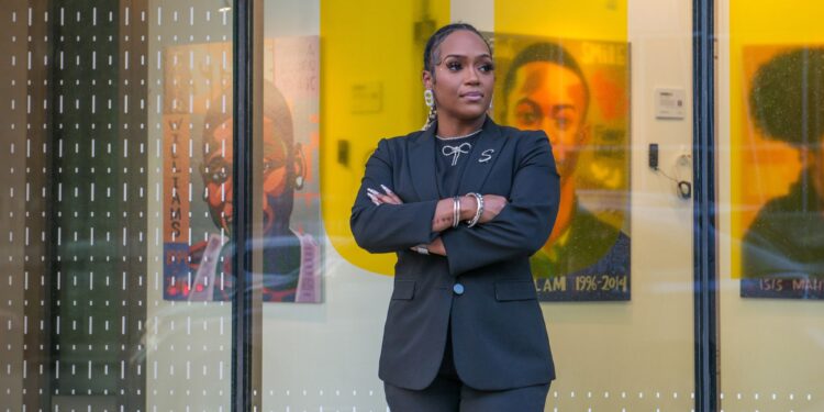 A photo of Shamari Jackson, a Black woman wearing a suit, standing outside of a building with glass windows. In the building space are paintings of homicide victims.