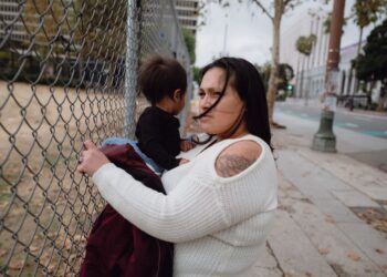 A photo shows Jazmine Mapes, a Native American woman with long dark hair, wearing a white sweater, holding onto a chain link fence with one hand and holding her infant son in the other.