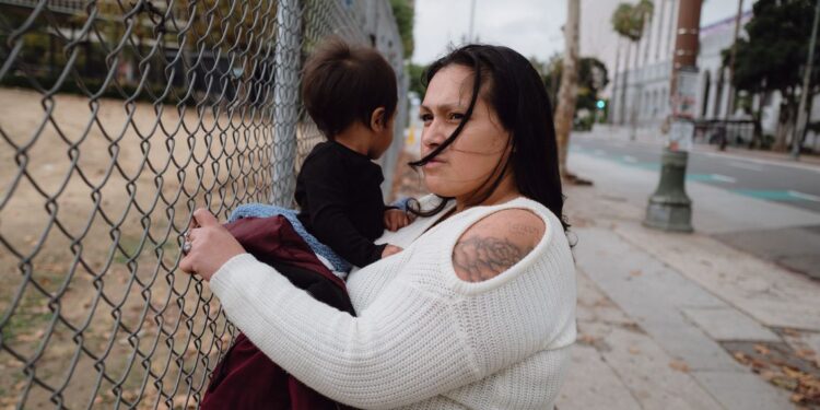 A photo shows Jazmine Mapes, a Native American woman with long dark hair, wearing a white sweater, holding onto a chain link fence with one hand and holding her infant son in the other.