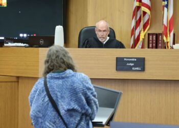 A balding White man wearing a black judge’s robe sits at the bench, while a person with gray hair and a blue jacket with their back to the camera makes their case to the judge.