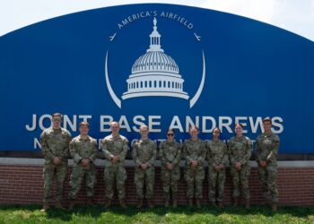 Eight USAFA cadets in uniform pose in front of a blue sign for Joint Base Andrews.