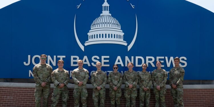 Eight USAFA cadets in uniform pose in front of a blue sign for Joint Base Andrews.