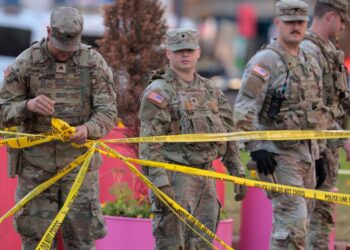 National Guard members respond to a shooting, with one soldier in the foreground holding yellow "POLICE LINE DO NOT CROSS" tape.