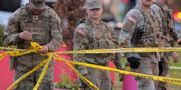 National Guard members respond to a shooting, with one soldier in the foreground holding yellow "POLICE LINE DO NOT CROSS" tape.