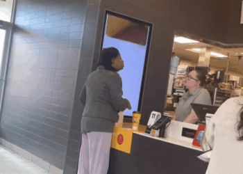 A woman in a gray jacket and light pants stands at a McDonald's counter speaking with an employee.