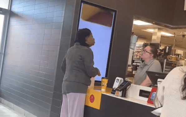 A woman in a gray jacket and light pants stands at a McDonald's counter speaking with an employee.