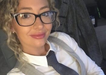 A woman with curly blonde hair and glasses wearing a white shirt, a gray vest, and a patterned blue tie.