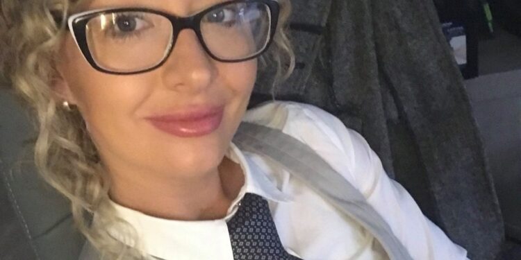 A woman with curly blonde hair and glasses wearing a white shirt, a gray vest, and a patterned blue tie.