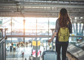 Female traveler with backpack and luggage looking out a window at an airport.
