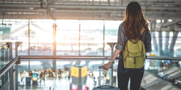 Female traveler with backpack and luggage looking out a window at an airport.