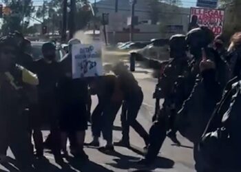 Federal officers pepper spray two individuals during a protest. There are more protesters marching in the background.