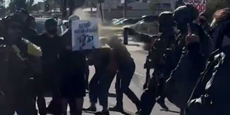 Federal officers pepper spray two individuals during a protest. There are more protesters marching in the background.