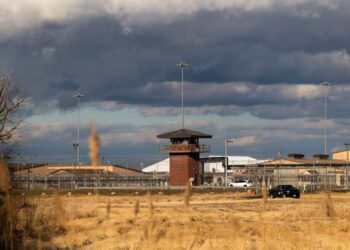 A photo shows a prison that has a red-brick lookout tower. Chain link fencing with barbed wire on top is behind the tower. In front of the tower is a field of yellow grass. The sky is mostly filled with dark clouds, with patches of pale blue.