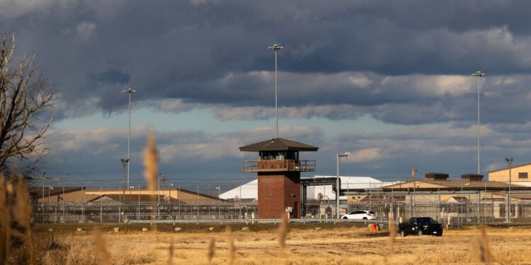 A photo shows a prison that has a red-brick lookout tower. Chain link fencing with barbed wire on top is behind the tower. In front of the tower is a field of yellow grass. The sky is mostly filled with dark clouds, with patches of pale blue.