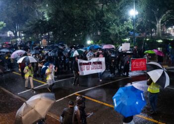 A group of protesters march in the street at night in the rain. People at the front of the group hold a banner that reads “ICE/Border Patrol out of NOLA.”