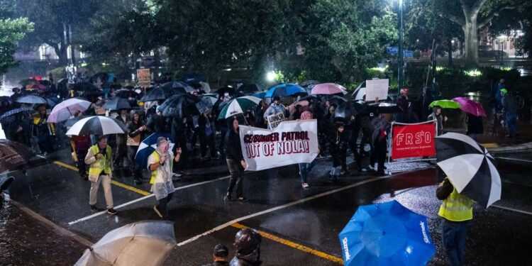A group of protesters march in the street at night in the rain. People at the front of the group hold a banner that reads “ICE/Border Patrol out of NOLA.”