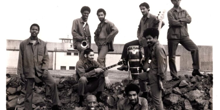 A photo of nine men wearing prison uniforms standing atop a pile of rocks at a prison. Some are standing at the ridge of the rockpile, one of them holding a saxophone and another, his left leg splayed over a bongo drum. In front of them sits a man with long sideburns and a guitar resting on his lap. On each side of him stand two men. In the foreground, one man with an afro sits smiling, his right elbow on his right knee. Just to the right sits a bald man, smiling, and holding a guitar.