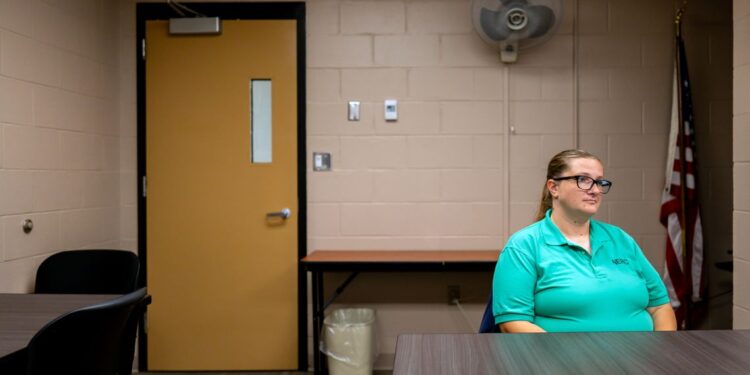 A photo shows Barbara Devine, a White woman with glasses wearing a green T-shirt that is part of a uniform, sitting in a room in front of a table. An American flag is visible behind her on the right, and a yellow door is visible behind her on the left.