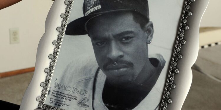 The hands of a Black woman hold the silver-colored framed black-and-white photo of her son, a young Black man wearing a dark-colored baseball cap with the logo of the Georgetown University Hoyas bulldog, a neatly trimmed goatee, a studded earring, and a light-colored baseball-style jersey.
