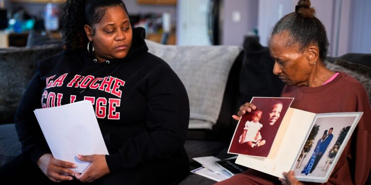 A photo shows Amber Hall, left, a Black woman wearing a black sweatshirt that reads “La Roche College” holding some white sheets of paper. Her mother, Cora Fleeta Hill, right, a Black woman wearing a maroon matching sweatshirt and sweatpants, holds some photos showing her son Jayson Murphy while they sit on a couch.