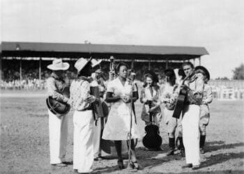 A black-and-white photo of a group of musicians dressed in white, with some wearing cowboy hats, standing in a horseshoe shape, while a Black woman in a white dress sings into a microphone. The group is standing on the rodeo grounds, and in the background are the stands where the audience is seated.